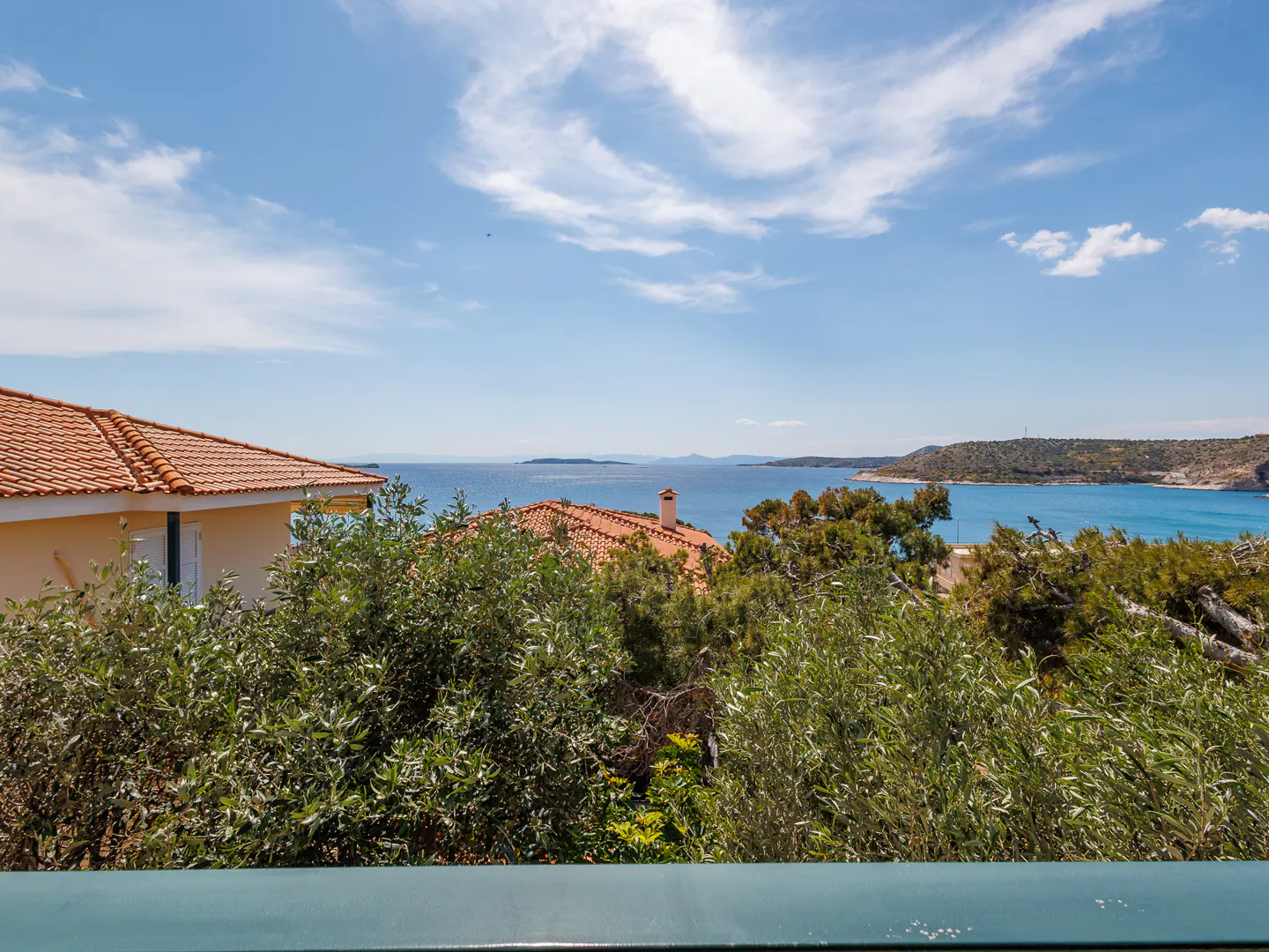 View of a blue ocean and sky from a balcony, with trees and houses with orange roofs in the foreground.