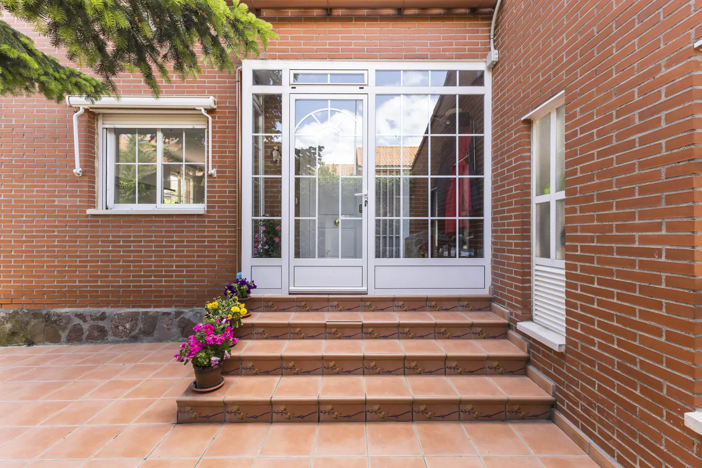 Brick house exterior with white framed glass door and windows. Terracotta tiled steps lead to the entrance, adorned with potted flowers.