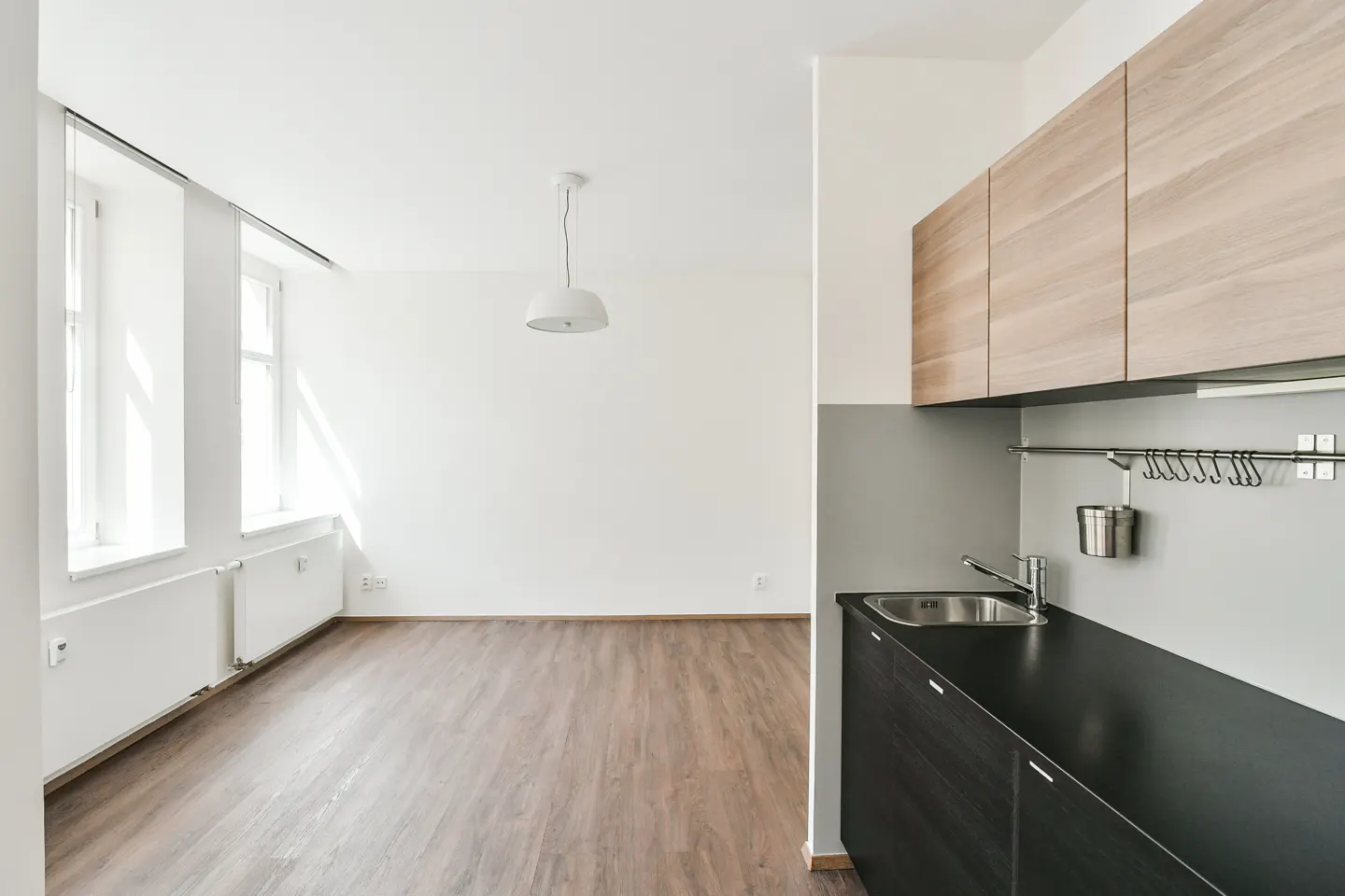 Modern kitchen with black counters and wood cabinets, opening into a bright, empty living area with wood floors and windows.