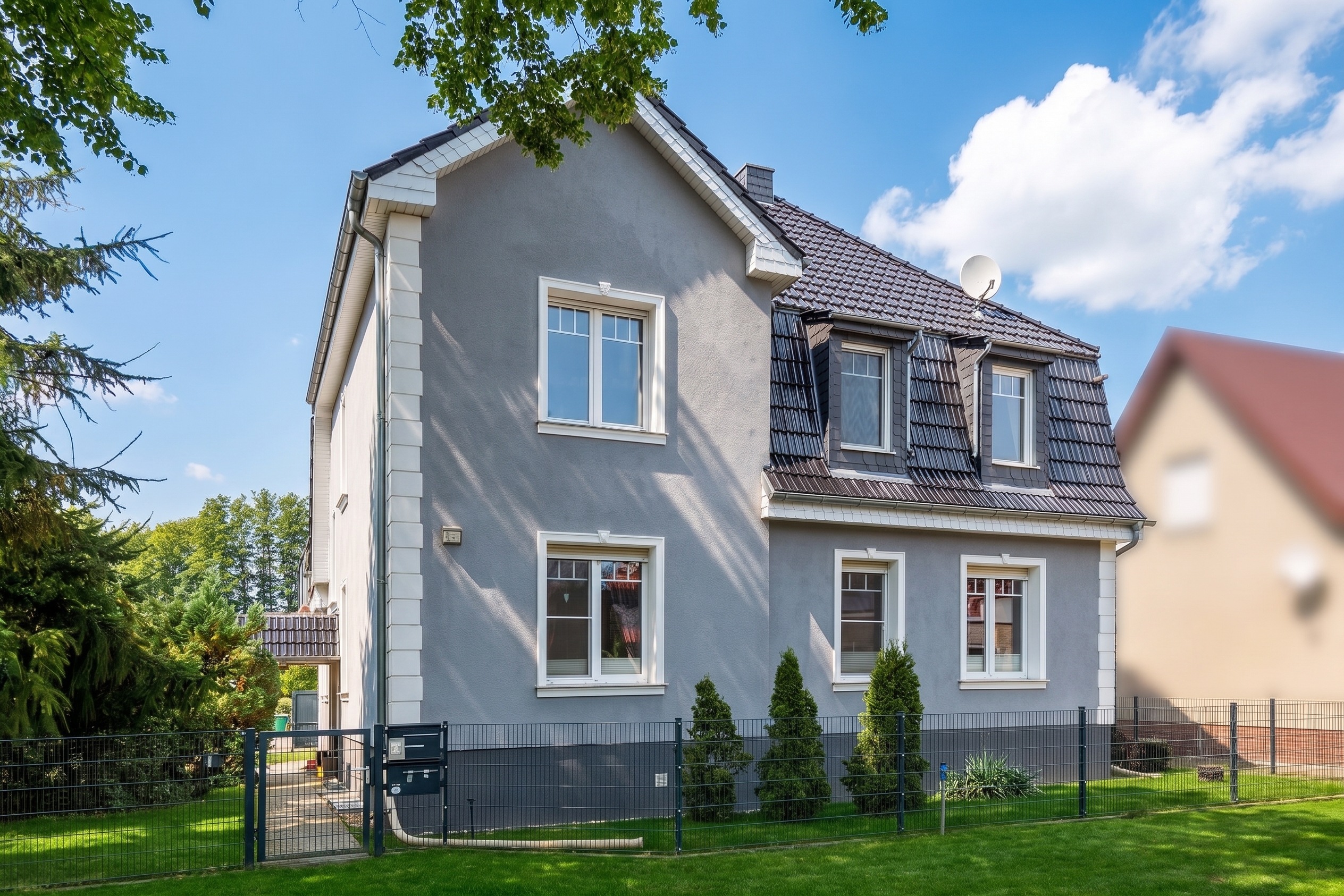 Two-story grey house with a dark tiled roof, white trim, and dormer windows, set behind a wire fence and green lawn.