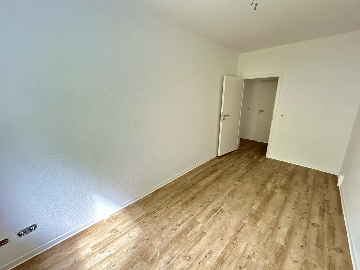 An empty room with white walls and wood-look flooring. An open white door leads to another room.