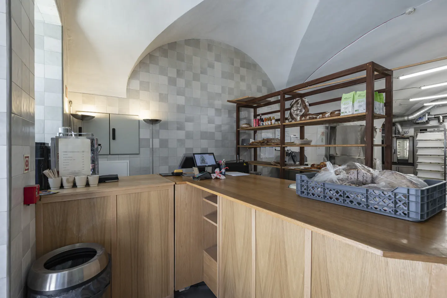 Bakery interior with a wooden counter, shelves with bread, and a tiled wall. A gray crate of bread sits on the counter.