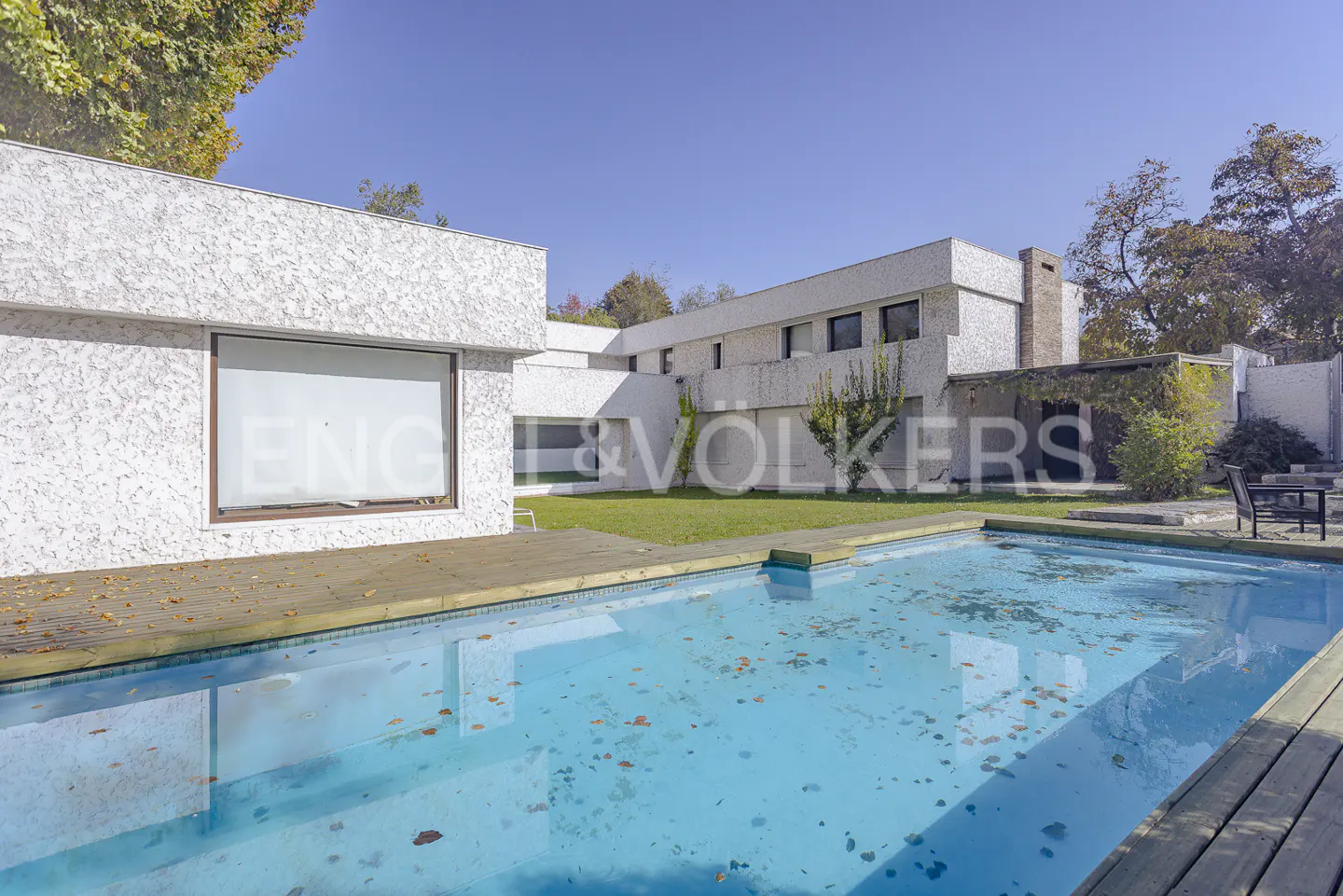 Modern white house with a swimming pool and wooden deck, surrounded by trees under a clear blue sky.