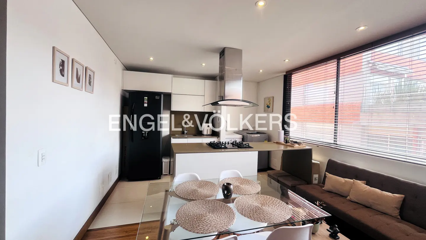 A modern apartment interior with a kitchen, dining area, and living space. White walls, a black refrigerator, and a glass dining table are visible.