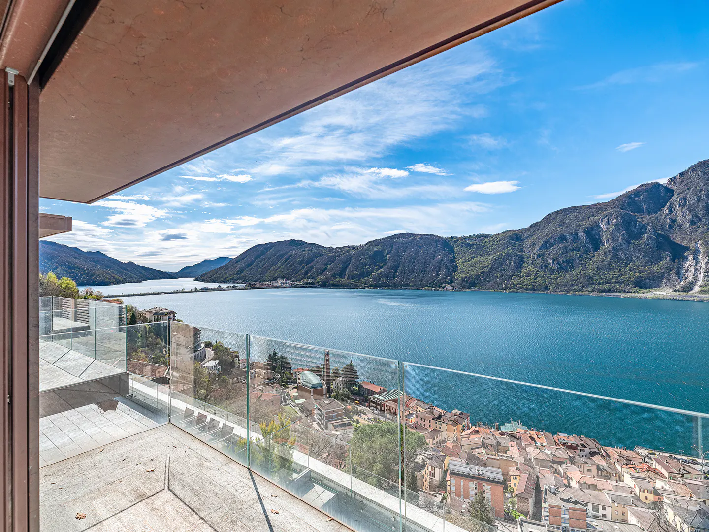 Balcony view of a blue lake and mountains under a blue sky. A town with red roofs sits below. Glass railing.
