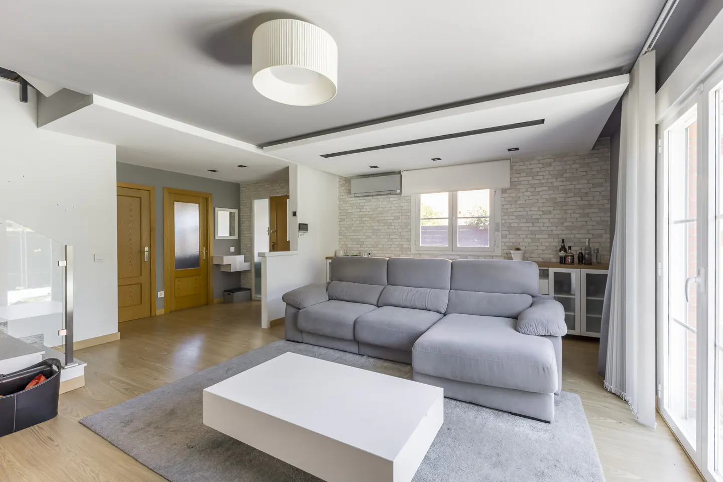 Living room with gray sofa, white coffee table on gray rug, and light wood floors. White brick wall and large windows.