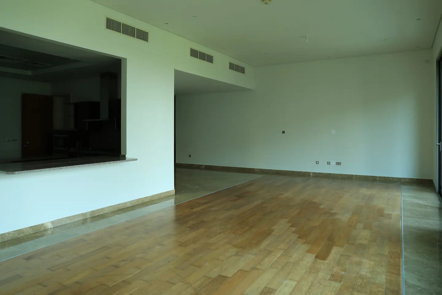 Empty room with light wood floors and white walls. A kitchen is visible through a serving hatch.