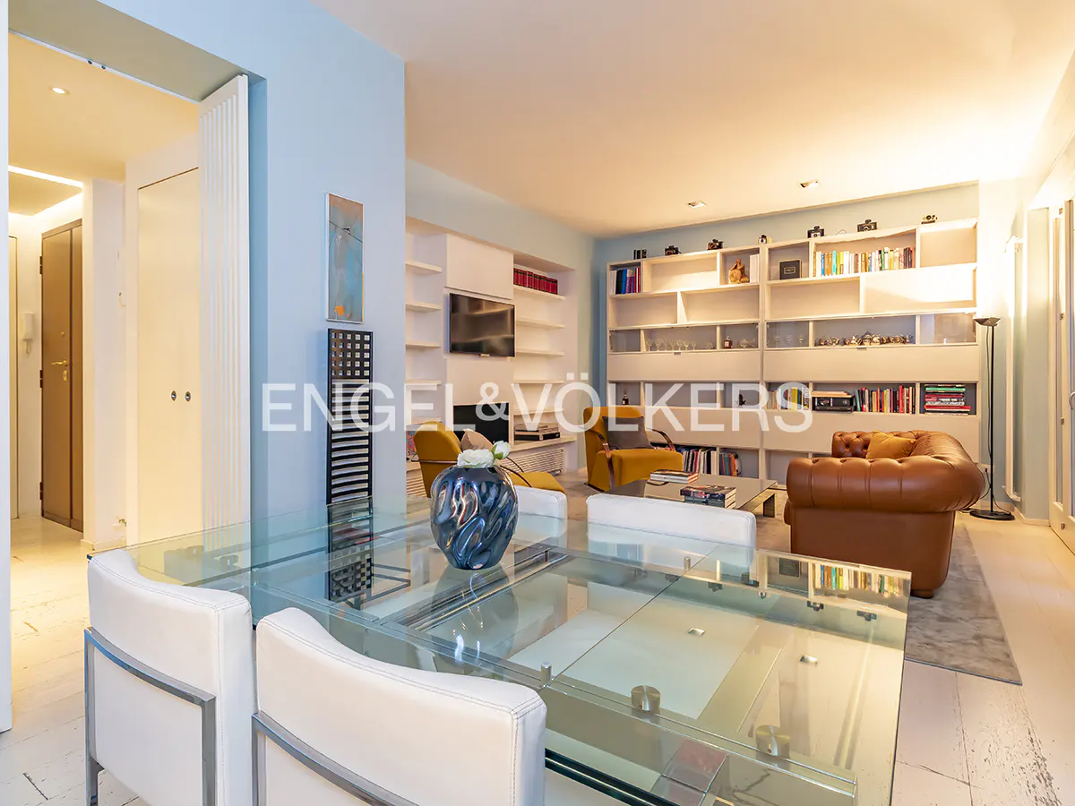 Bright living room with glass table, white chairs, and built-in bookshelves filled with books and decorative items. Yellow armchairs and a brown leather sofa add warmth.
