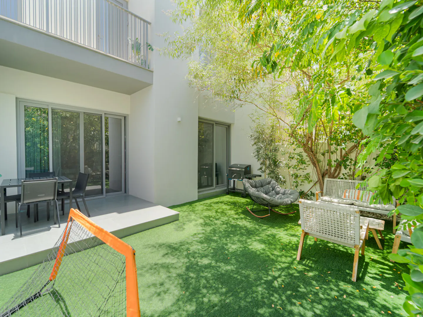 Modern backyard patio with artificial turf, outdoor seating, a grill, and a small soccer goal near a white house.