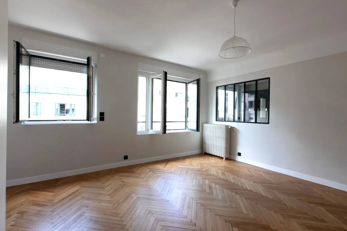 Empty room with light gray walls, herringbone wood floors, open windows, a white radiator, and a black-framed interior window.