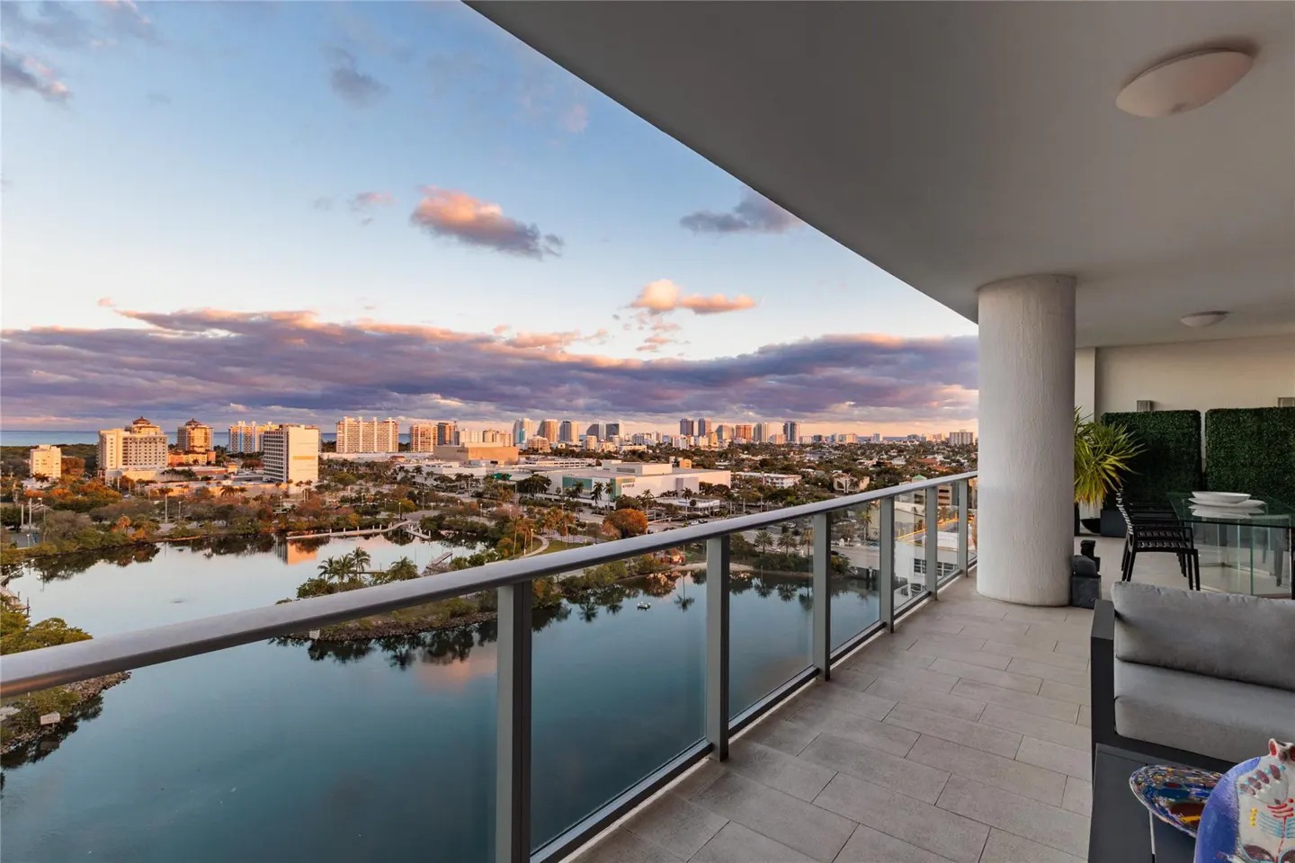 Balcony view of a city skyline at sunset. Glass railings overlook a lake reflecting the buildings and colorful sky. Outdoor furniture is visible.