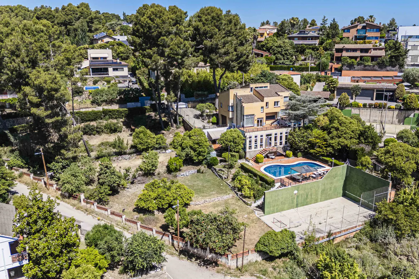 Aerial view of a yellow two-story house with a pool, tennis court, and lush green trees.