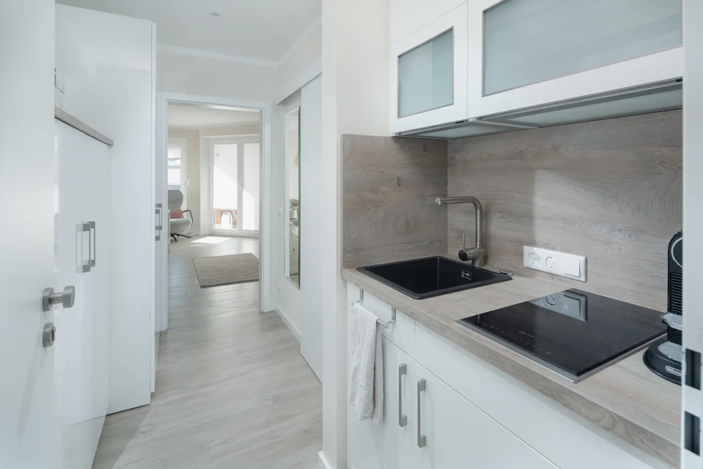 A bright, modern kitchen with white cabinets, a black sink, and a black stovetop. A hallway leads to a sunlit room with a chair and balcony.