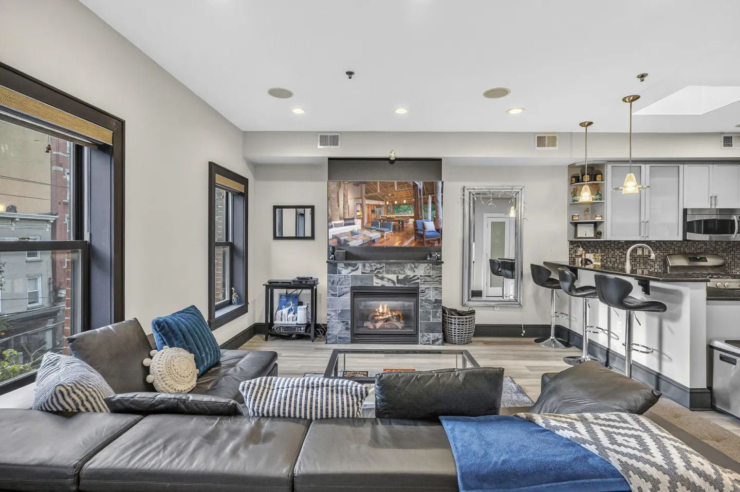 Modern living room with a black leather sectional, stone fireplace with a TV, and a kitchen island with bar stools.