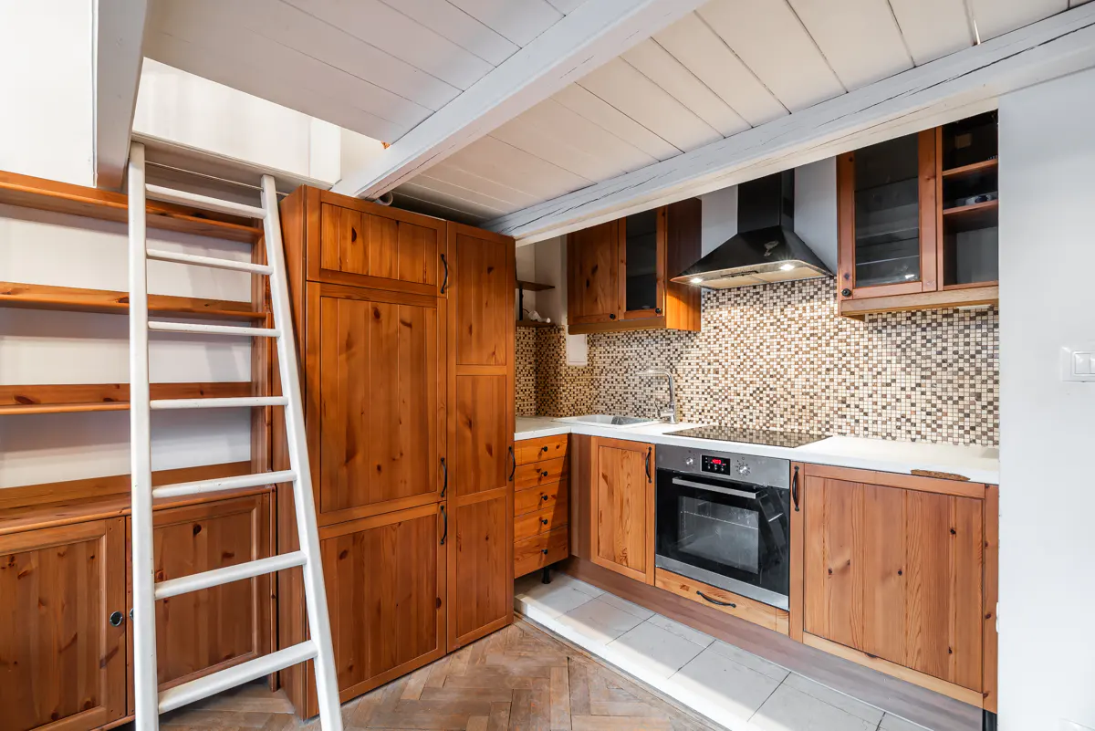 A compact kitchen with wooden cabinets, stainless steel appliances, and a white ladder leading to a loft area.