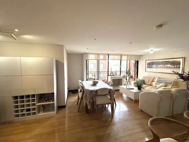Bright living room with hardwood floors, white walls, and large windows. A dining table with chairs sits near a balcony, and a white sofa faces a painting.