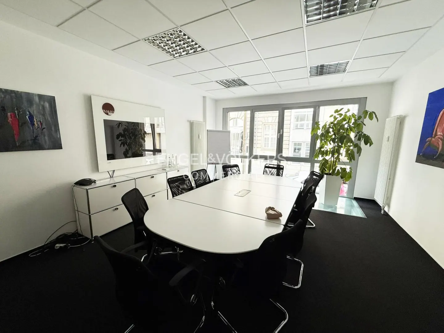 Bright conference room with a white table, black chairs, and black carpet. A large window lets in natural light. Artwork adorns the white walls.