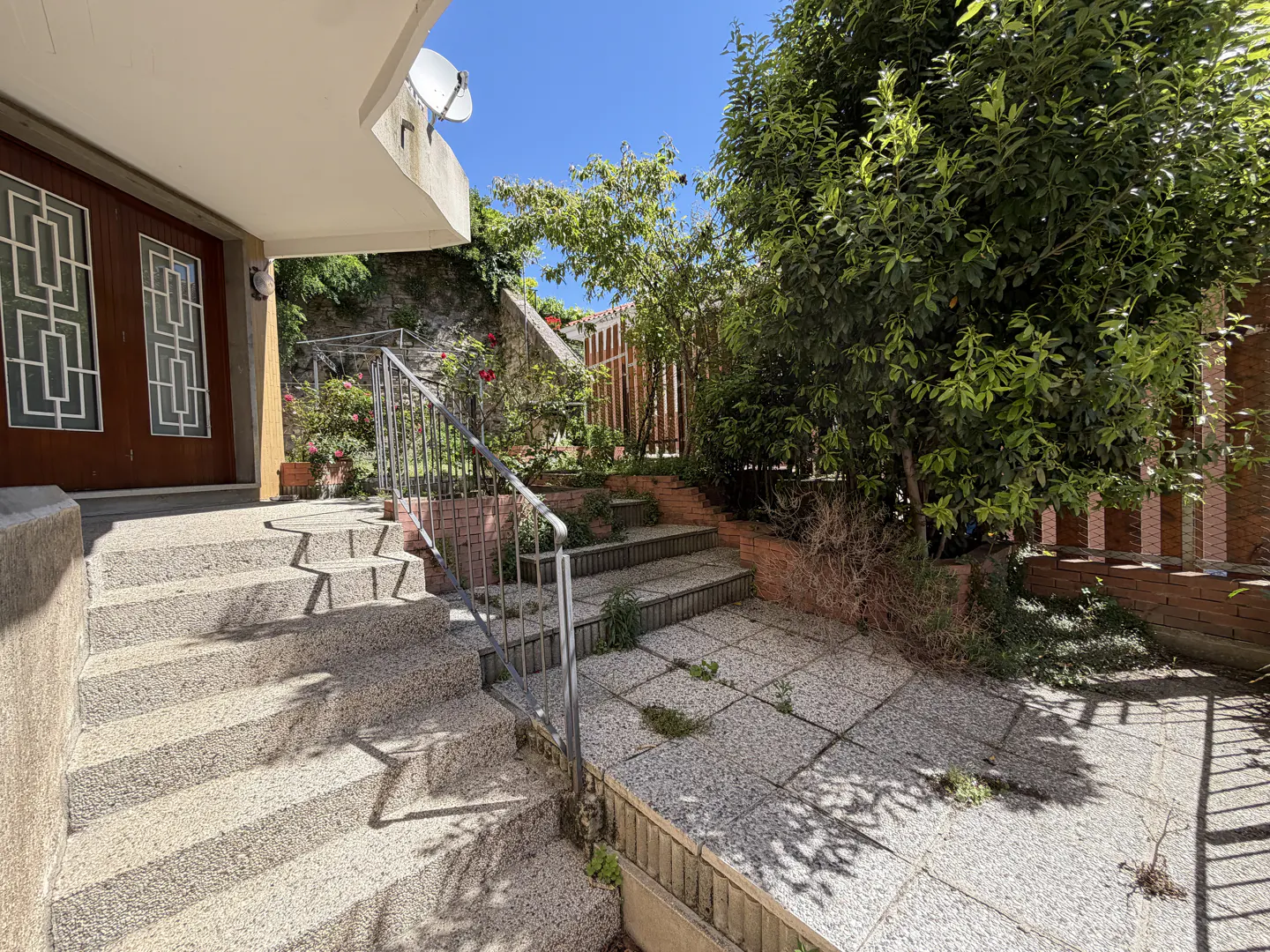 Exterior view of a house with stone steps leading to a brown front door, a satellite dish, and a garden with greenery.