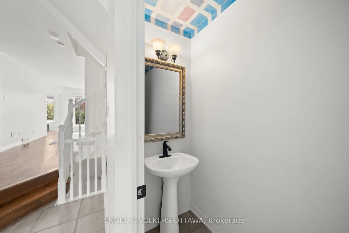 A powder room with a white pedestal sink, gold-framed mirror, and blue geometric ceiling design. A staircase is visible in the background.