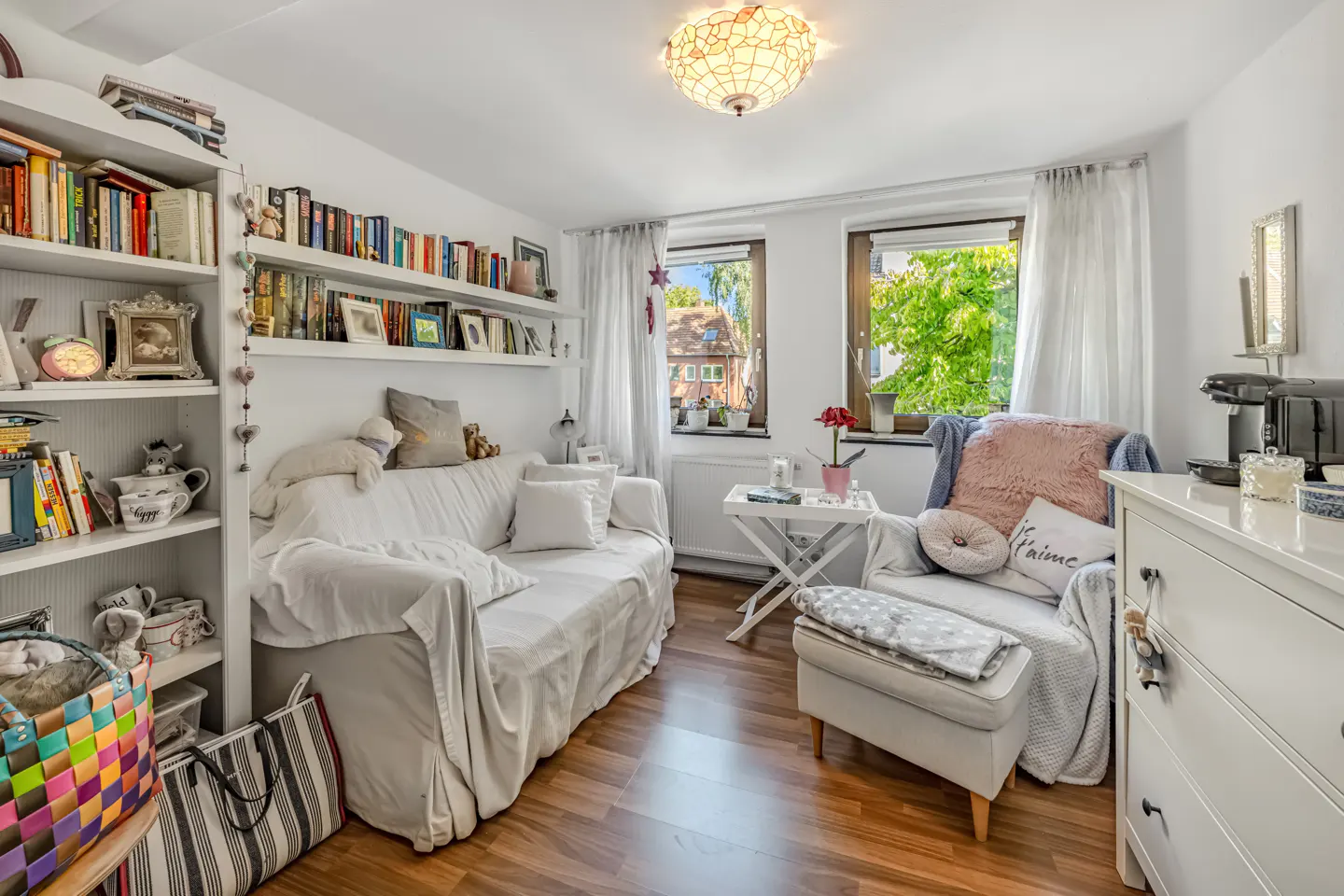 Cozy white living room with a bookshelf, sofa, chair, and wood floors. Windows show green trees outside.