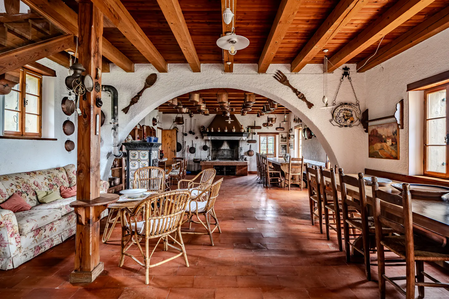 Rustic dining room with terracotta floors, wooden beams, and white walls. Wicker chairs surround tables, and copper pots hang above a large fireplace.