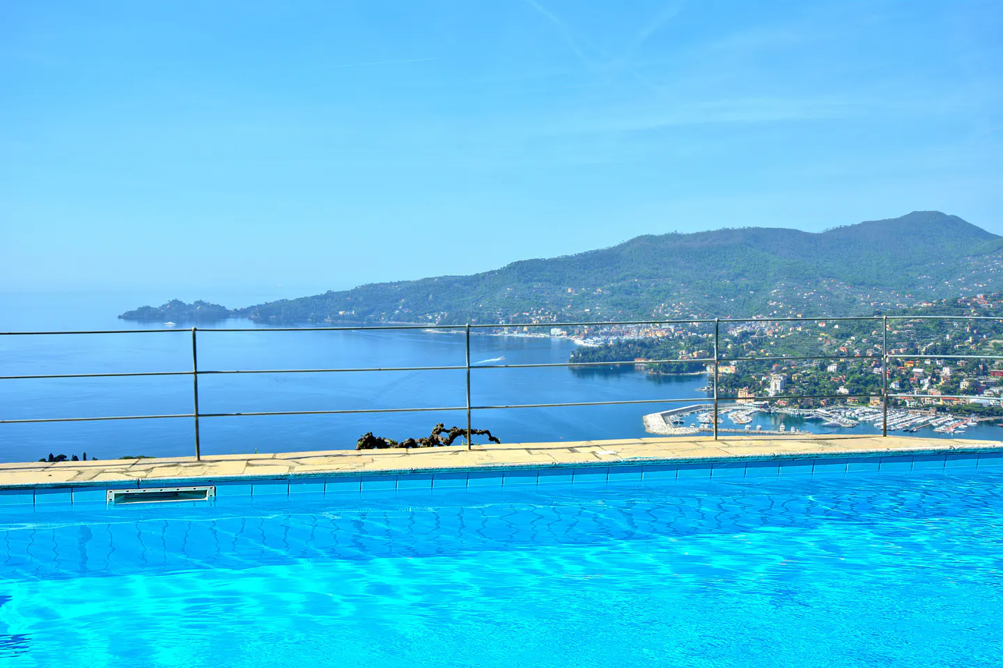 A bright blue pool overlooks a bay with mountains in the background under a clear blue sky.
