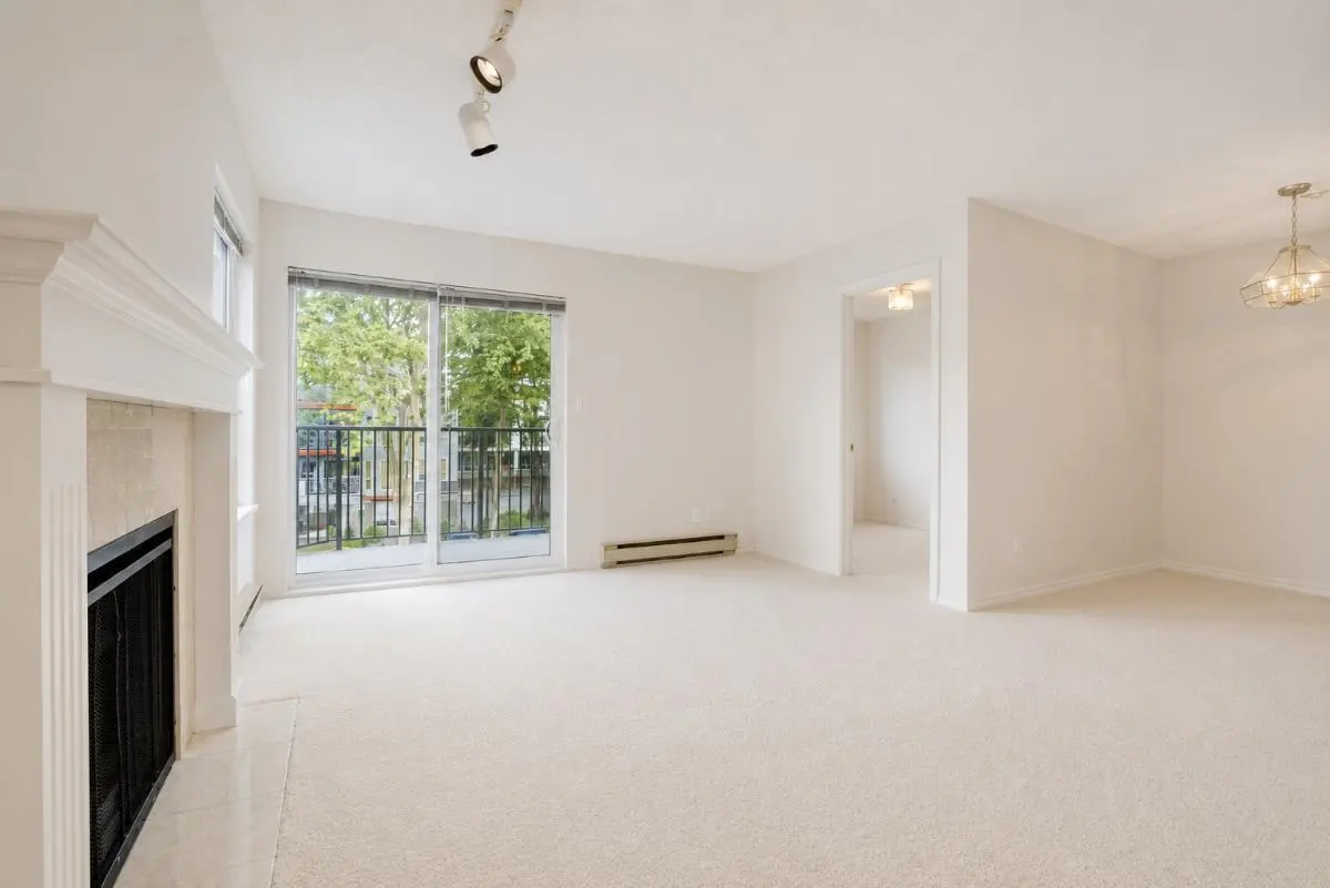 Bright, empty living room with beige carpet, a white fireplace, sliding glass doors to a balcony, and a chandelier.