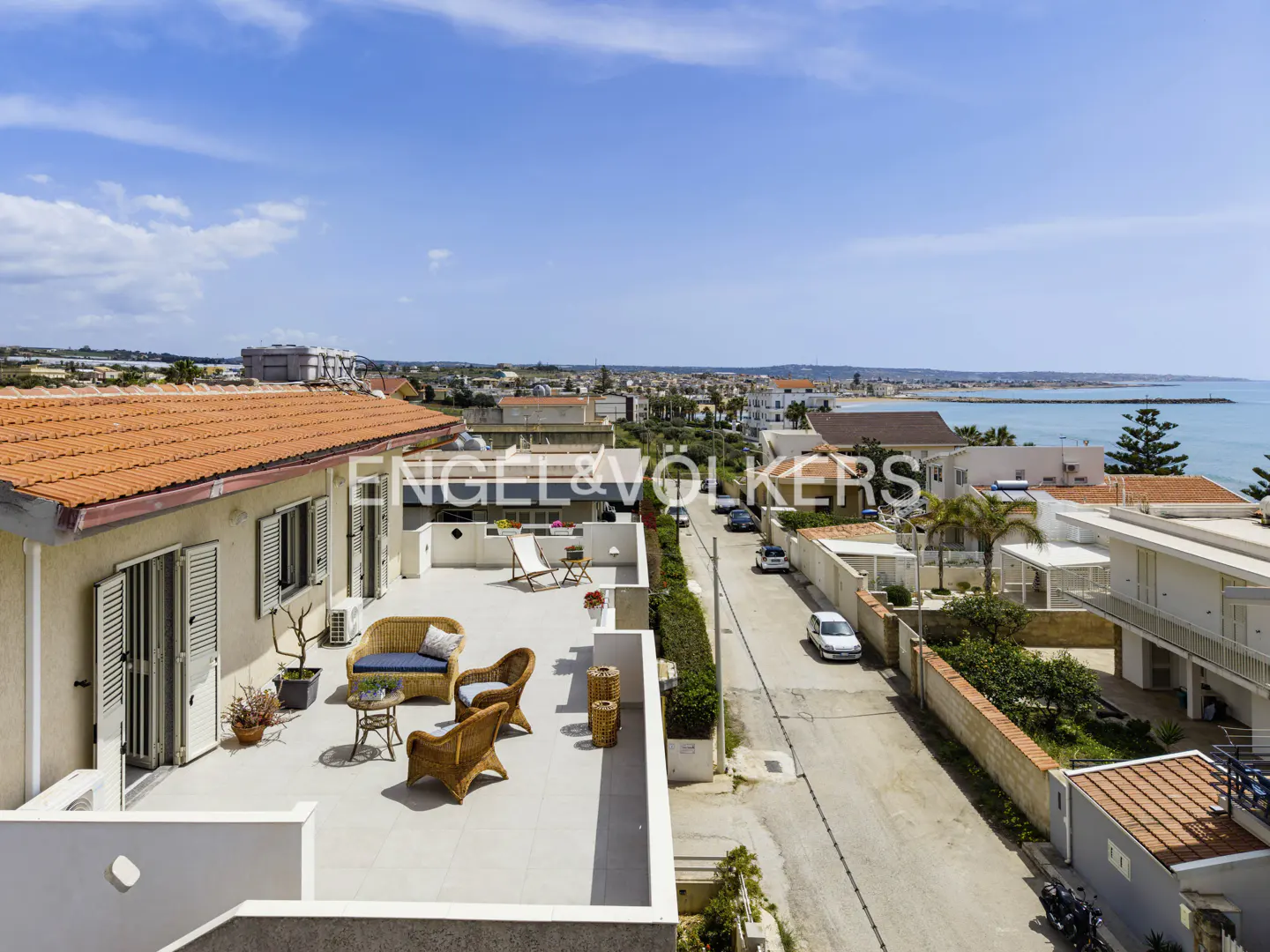Aerial view of a rooftop terrace with wicker furniture, overlooking a street, houses, and the ocean under a blue sky.
