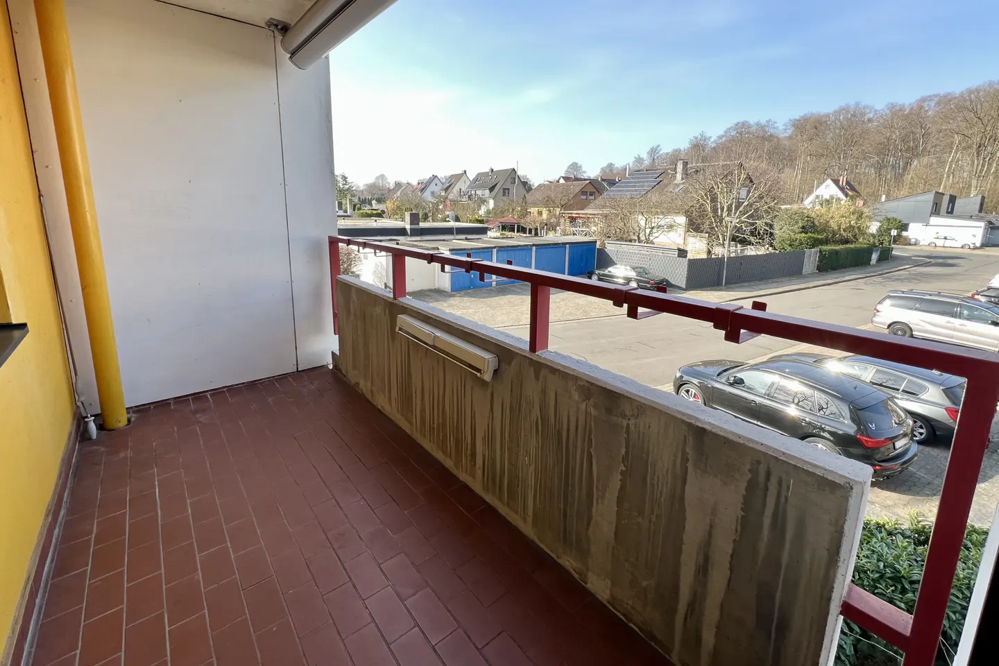 Balcony view with red brick floor, concrete railing, and red metal bars. Cars and houses are visible in the background.
