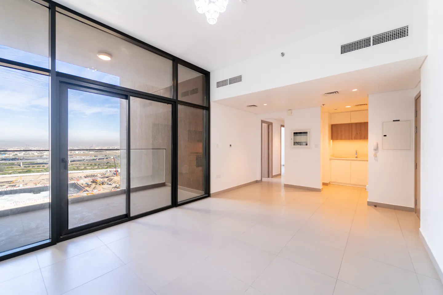 Bright, empty apartment with white walls, tile floor, and black-framed sliding glass doors to a balcony overlooking a city.