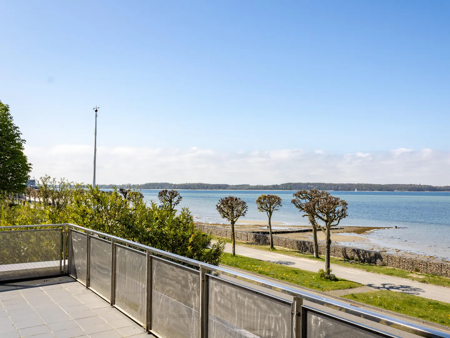 View from a balcony with a metal railing overlooking a blue bay with trees and a walking path.