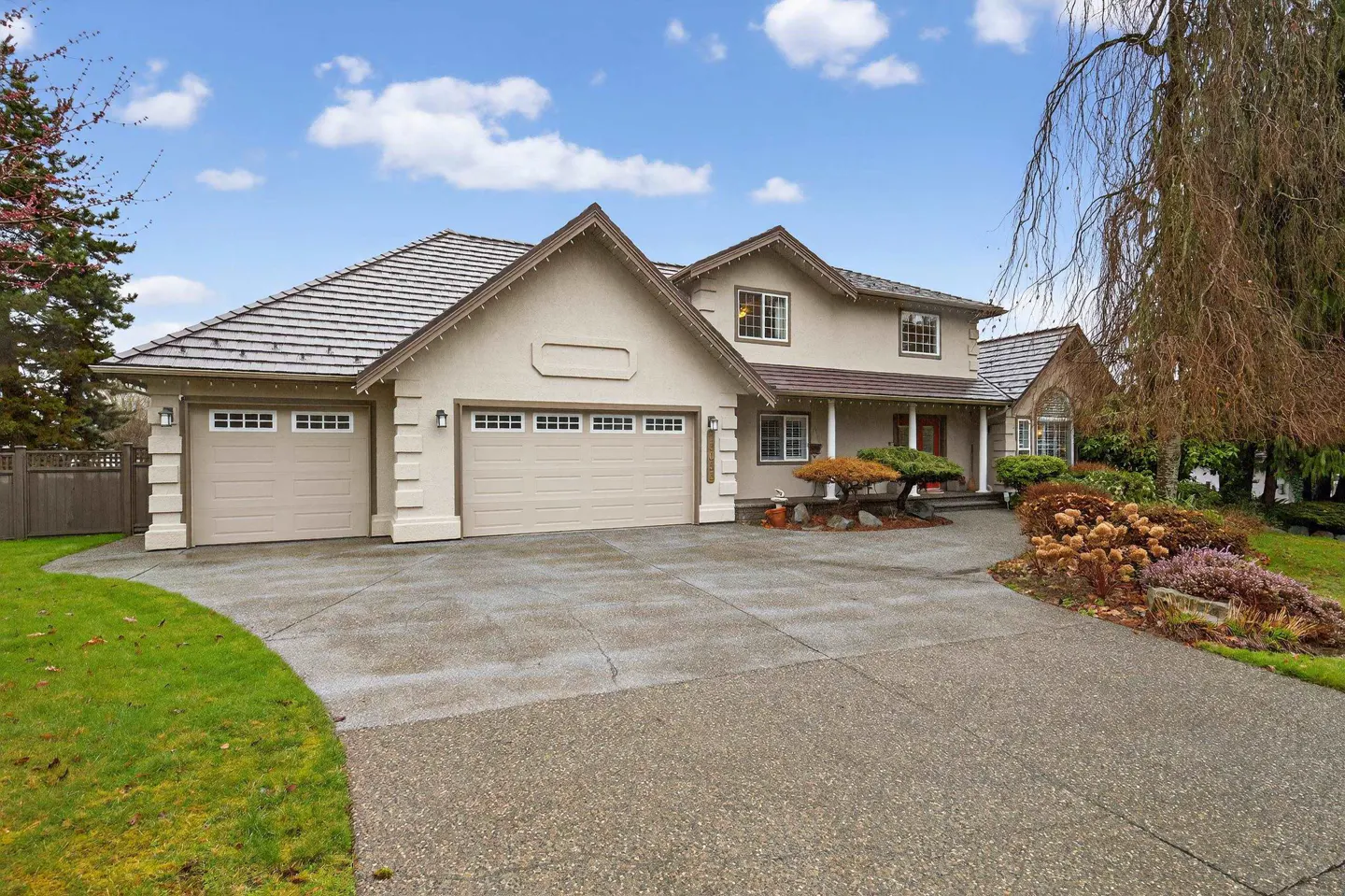 Two-story beige suburban home with a three-car garage, concrete driveway, and landscaped front yard under a blue sky.