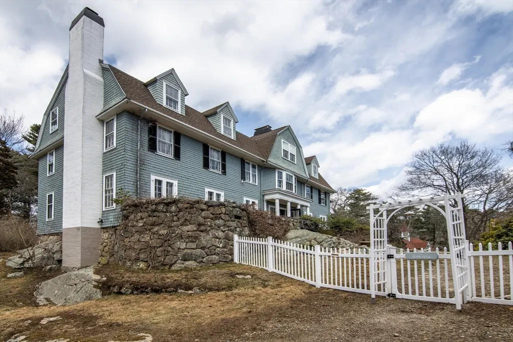 Exterior view of a large, light blue house with a white chimney, stone wall, and white picket fence.