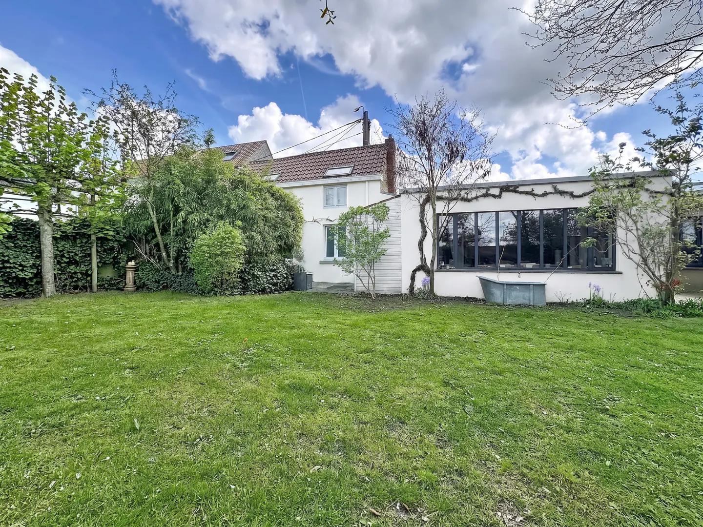 A wide shot of a house's backyard with a green lawn, trees, and a white house with a red roof under a cloudy sky.