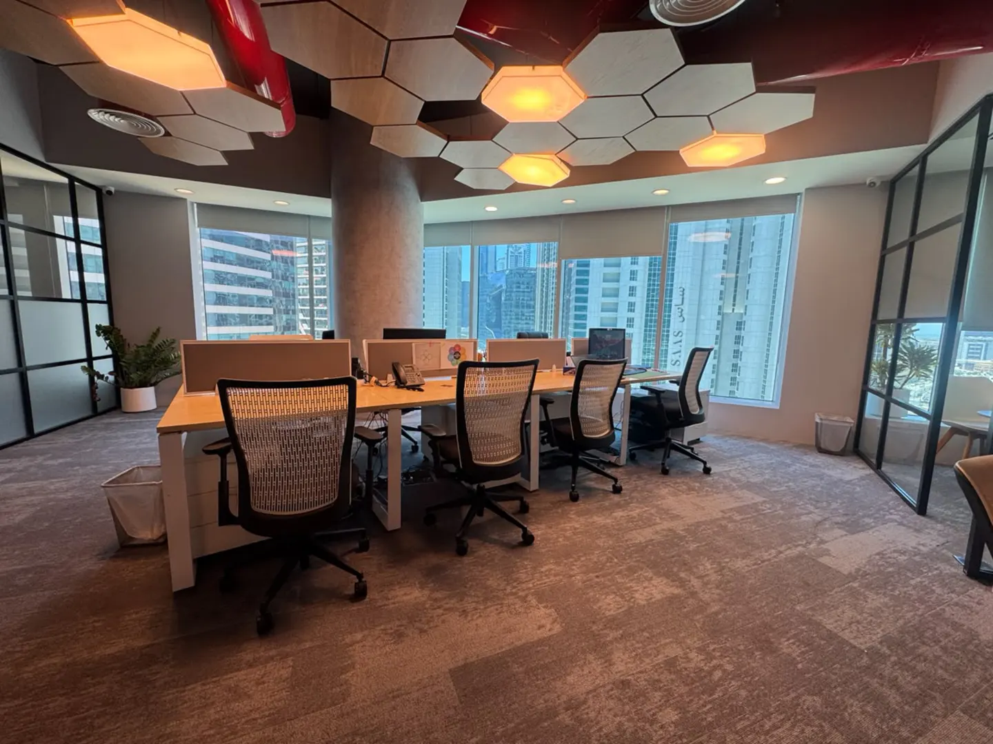 Modern office with desks, mesh chairs, and hexagonal ceiling lights, featuring large windows with city views.