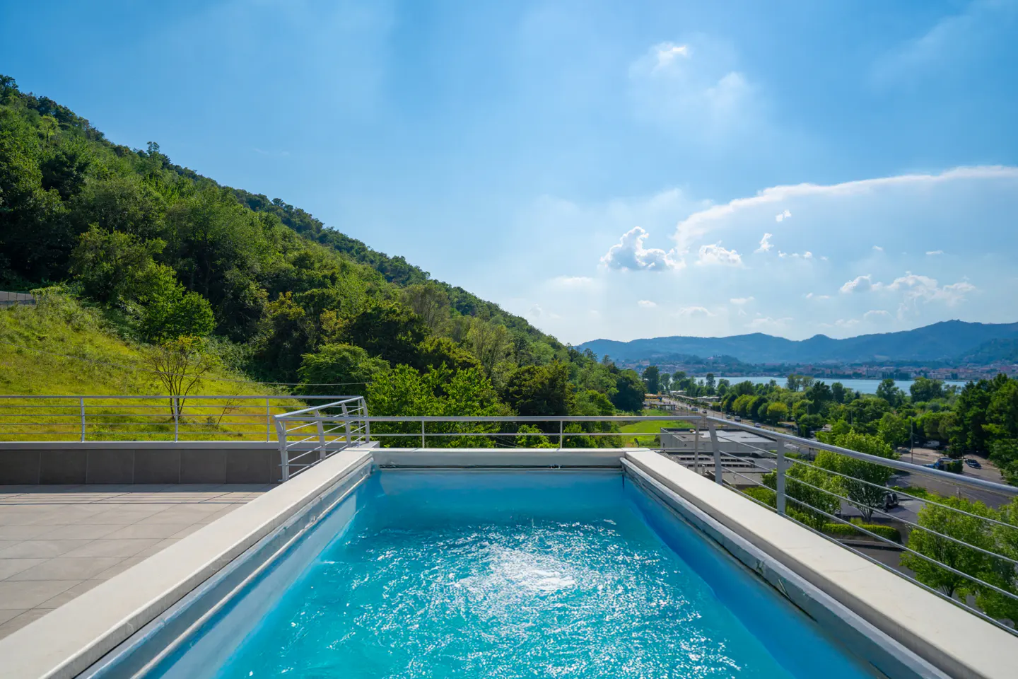 Rooftop pool with turquoise water, surrounded by a white deck and railing. Green hills and a blue sky with clouds in the background.