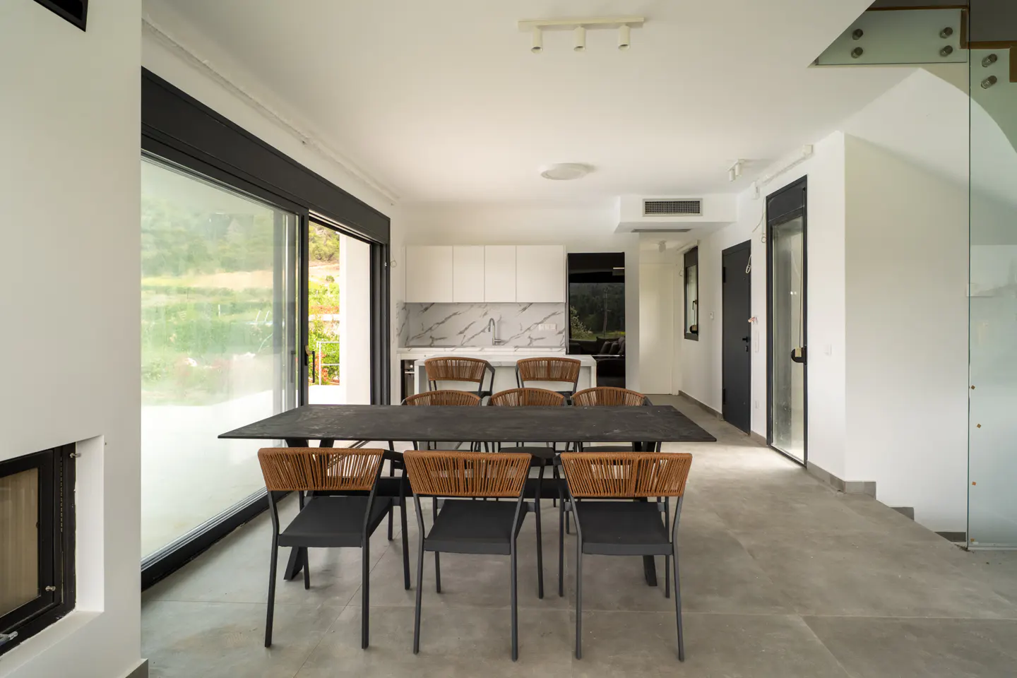 Bright, modern dining area with a long, dark table and woven-back chairs. White kitchen in the background, large window to the left.