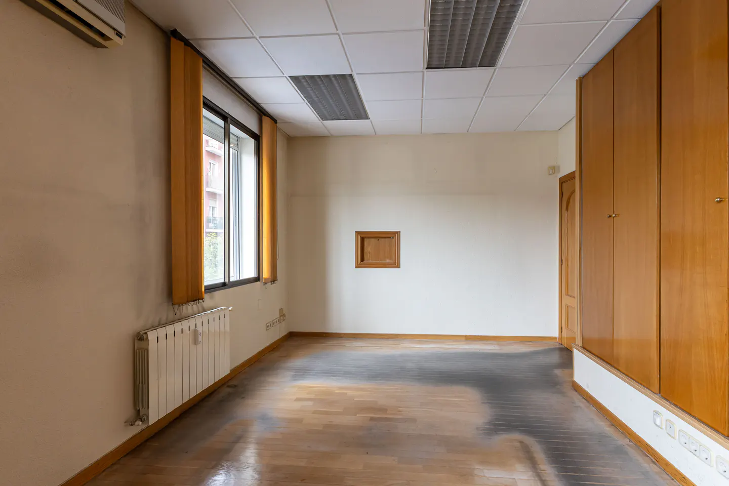 Empty room with wood floors, beige walls, and a white tile ceiling. A window with yellow curtains and a radiator are on the left. A wood cabinet is on the right.