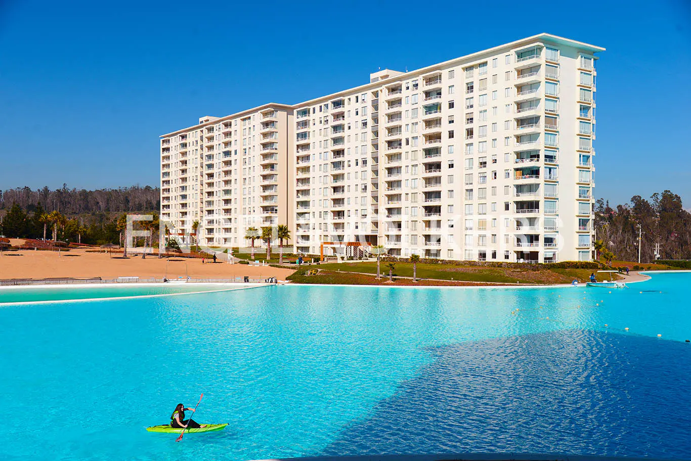 A tall, white apartment building overlooks a turquoise lagoon. A woman kayaks on the water. A sandy beach and trees are in the background.