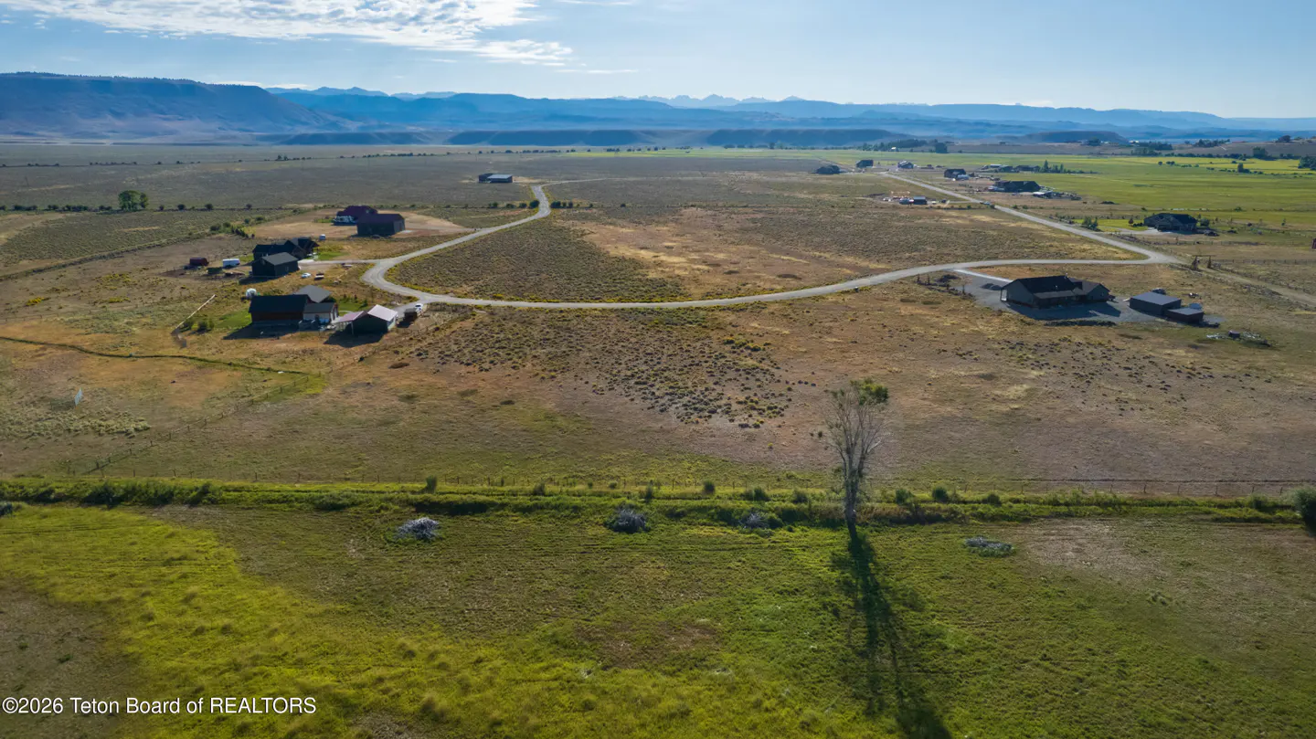 Aerial view of a rural landscape with scattered houses, dirt roads, and distant mountains under a blue sky.