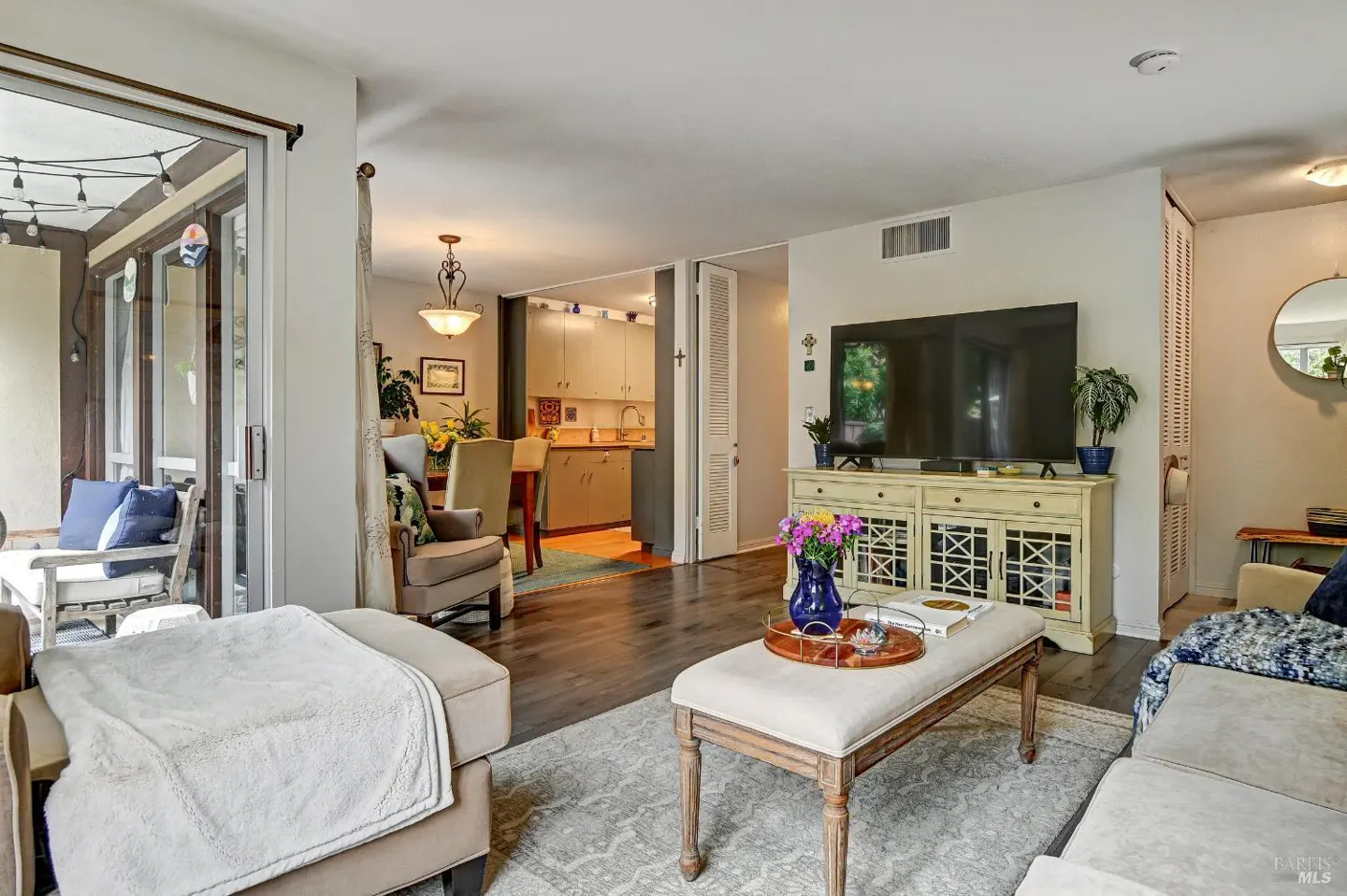 A bright living room with a beige sofa, ottoman, and a TV on a white cabinet. A dining area and kitchen are visible in the background.