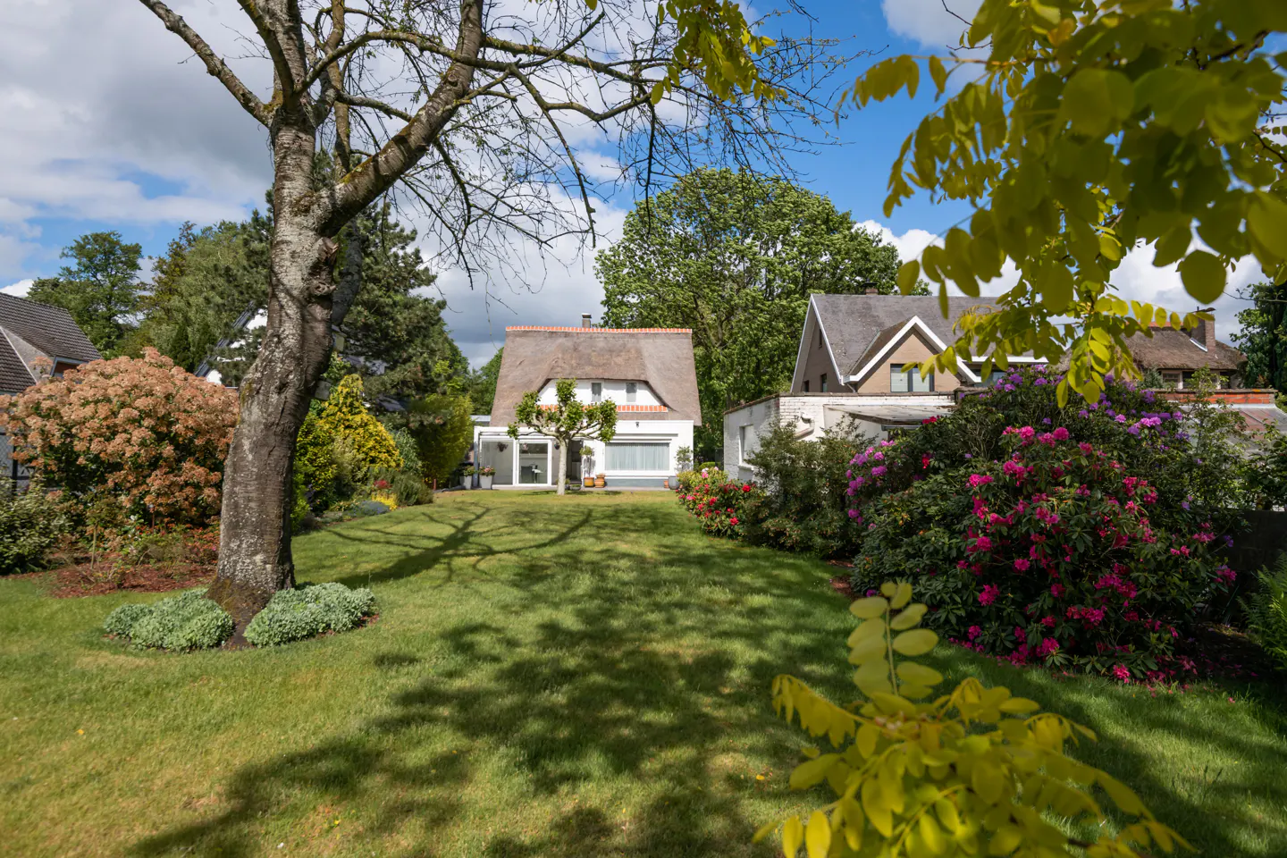 A white house with a thatched roof sits on a green lawn surrounded by trees and flowering bushes.