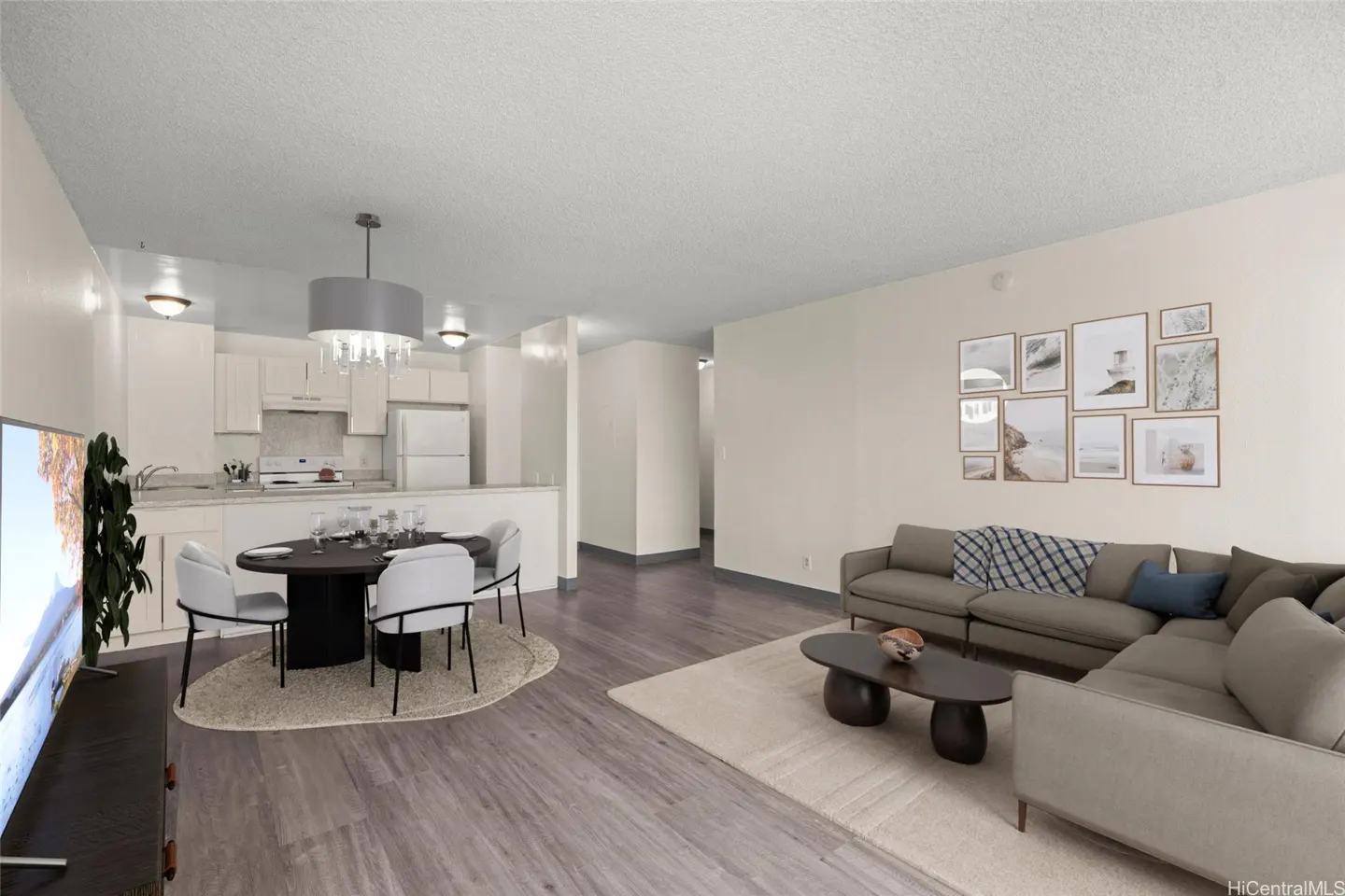 Open-concept living space with a gray sectional sofa, coffee table, and a gallery wall. A dining table and kitchen are in the background.