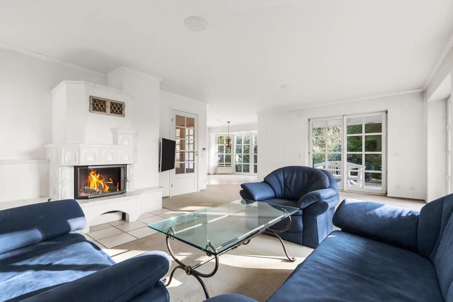 Bright living room with white walls, blue sofas, glass table, and a white fireplace with a burning fire.