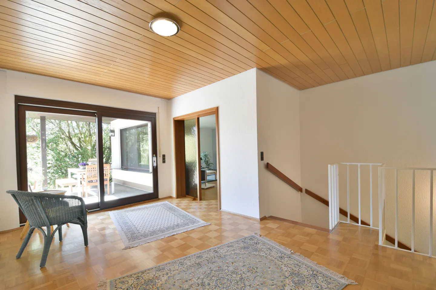 A bright foyer with a wood-paneled ceiling, sliding glass doors to a patio, and a staircase with white railings. A wicker chair and rugs accent the parquet floor.
