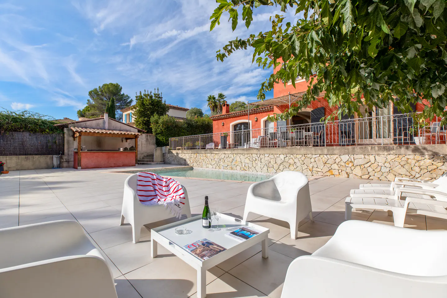 Outdoor patio with white chairs, table, and lounge chairs near a pool. A red house is in the background.