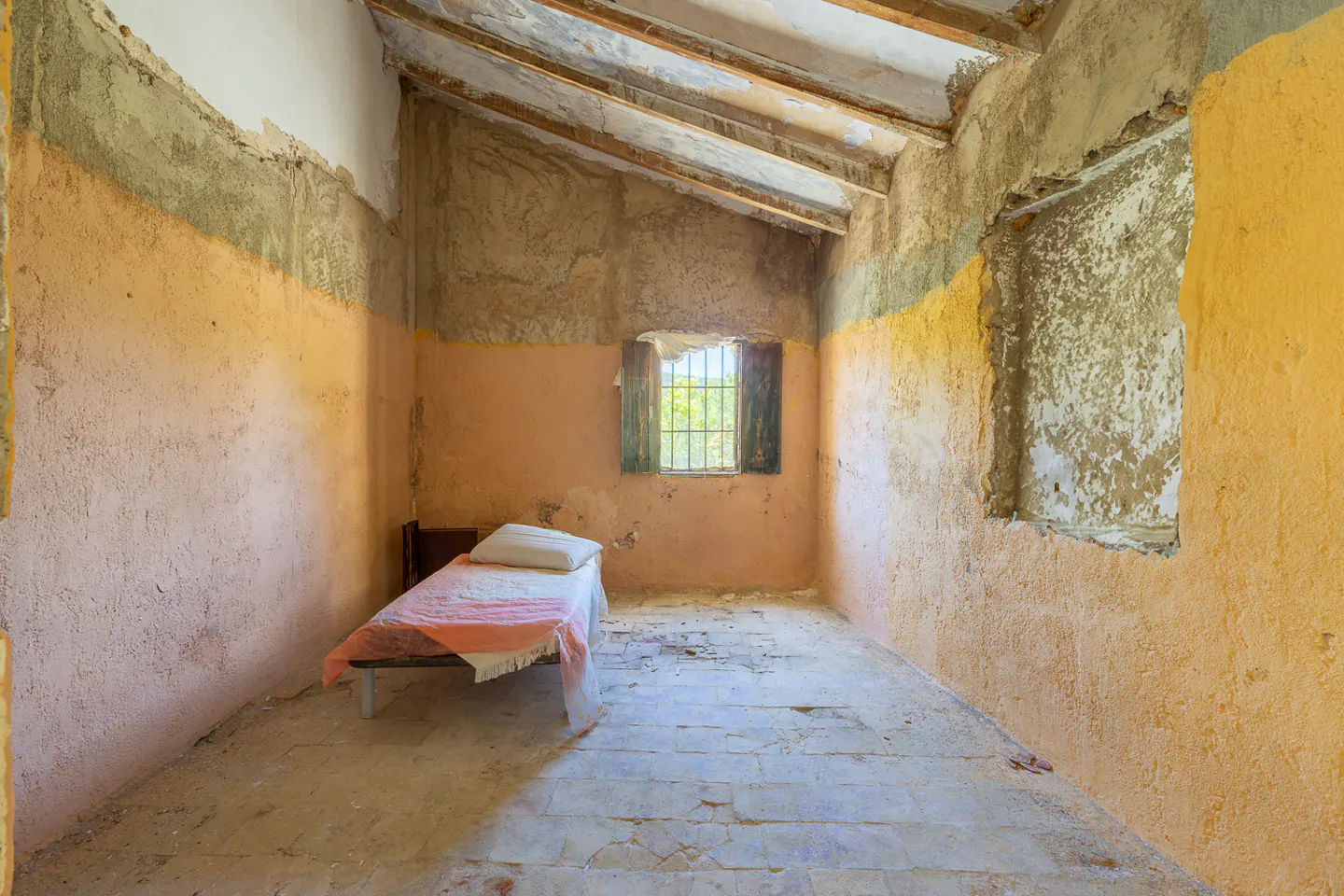 Interior of a dilapidated room with a single bed, barred window, and crumbling walls painted in peach and yellow.