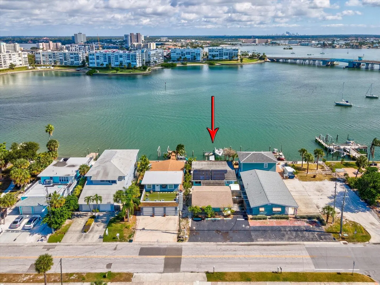 Aerial view of waterfront homes with docks, boats, and a red arrow pointing to a house with solar panels. City skyline in background.