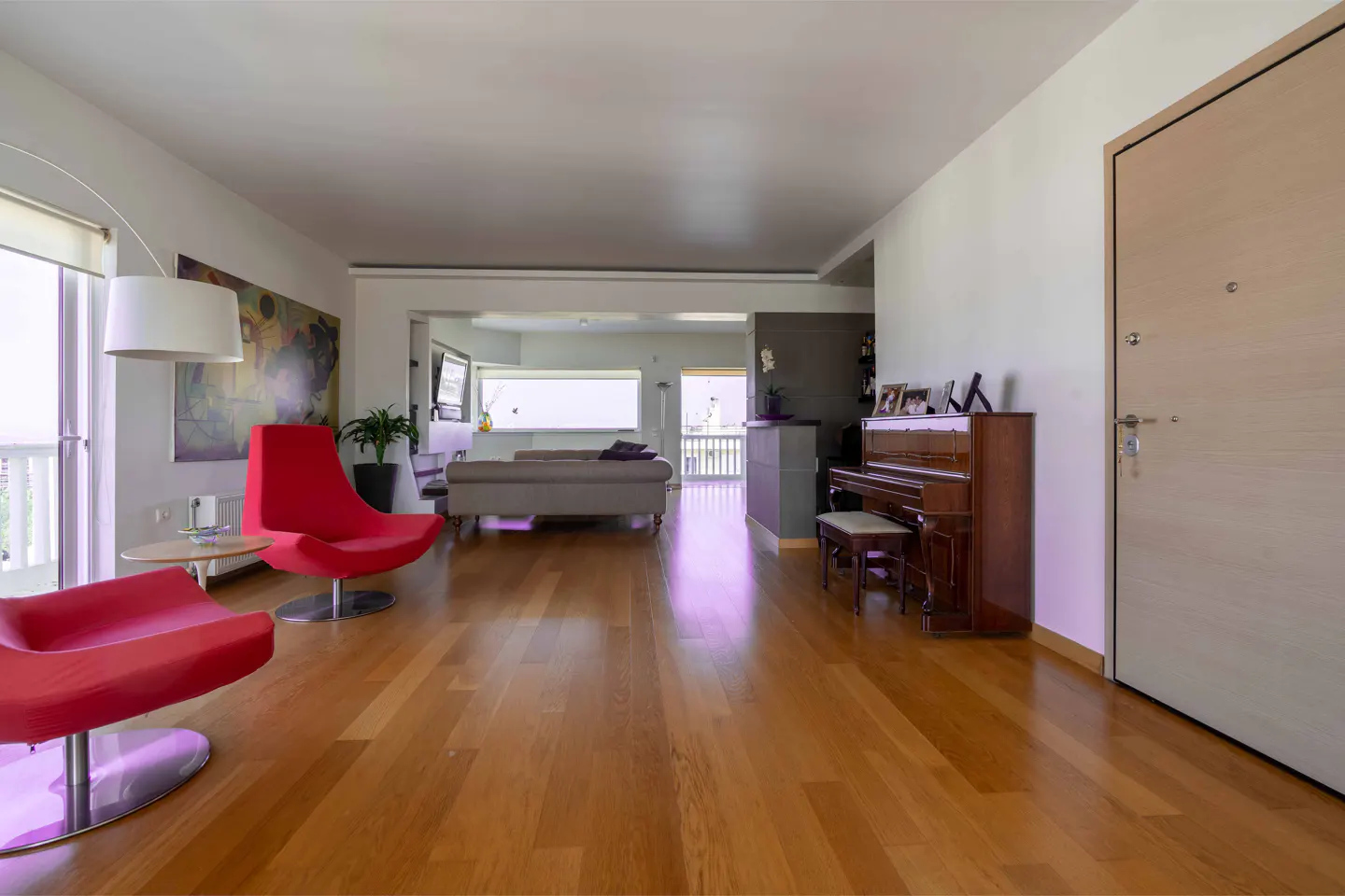 Bright, open living room with hardwood floors, red chairs, a piano, and a view to another room with a couch and balcony.