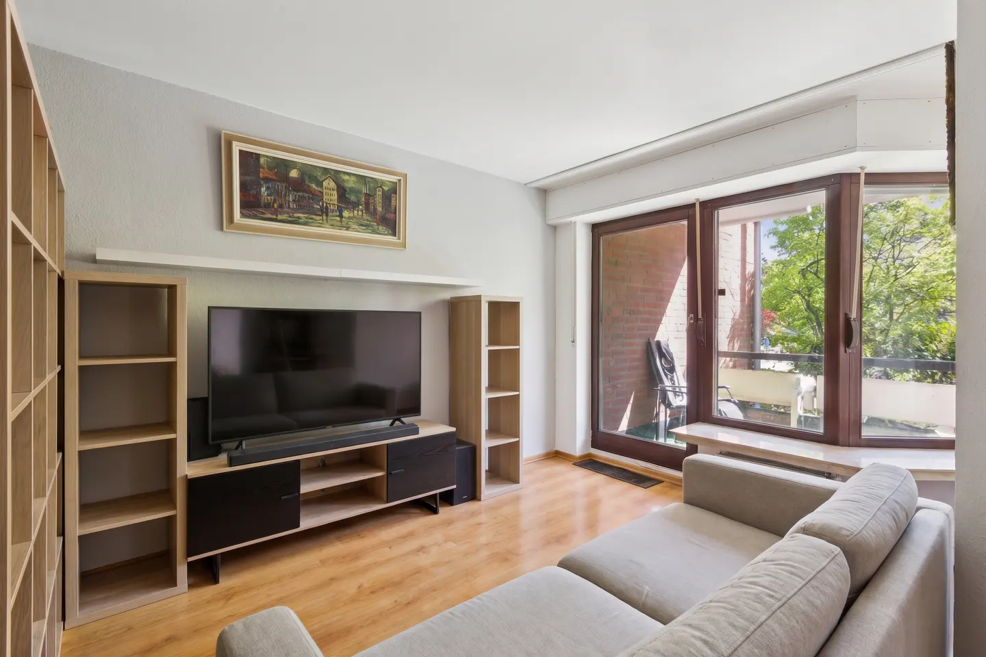 Living room with a gray sofa, a TV on a black and wood stand, and a balcony with a view of green trees.